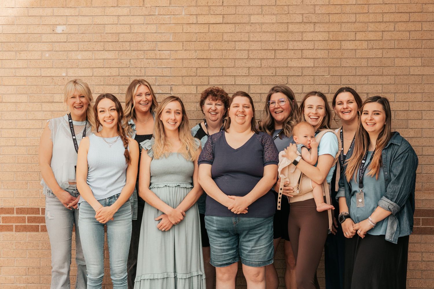 Group of ten smiling women posing together in front of a brick wall in casual and semi-formal attire.