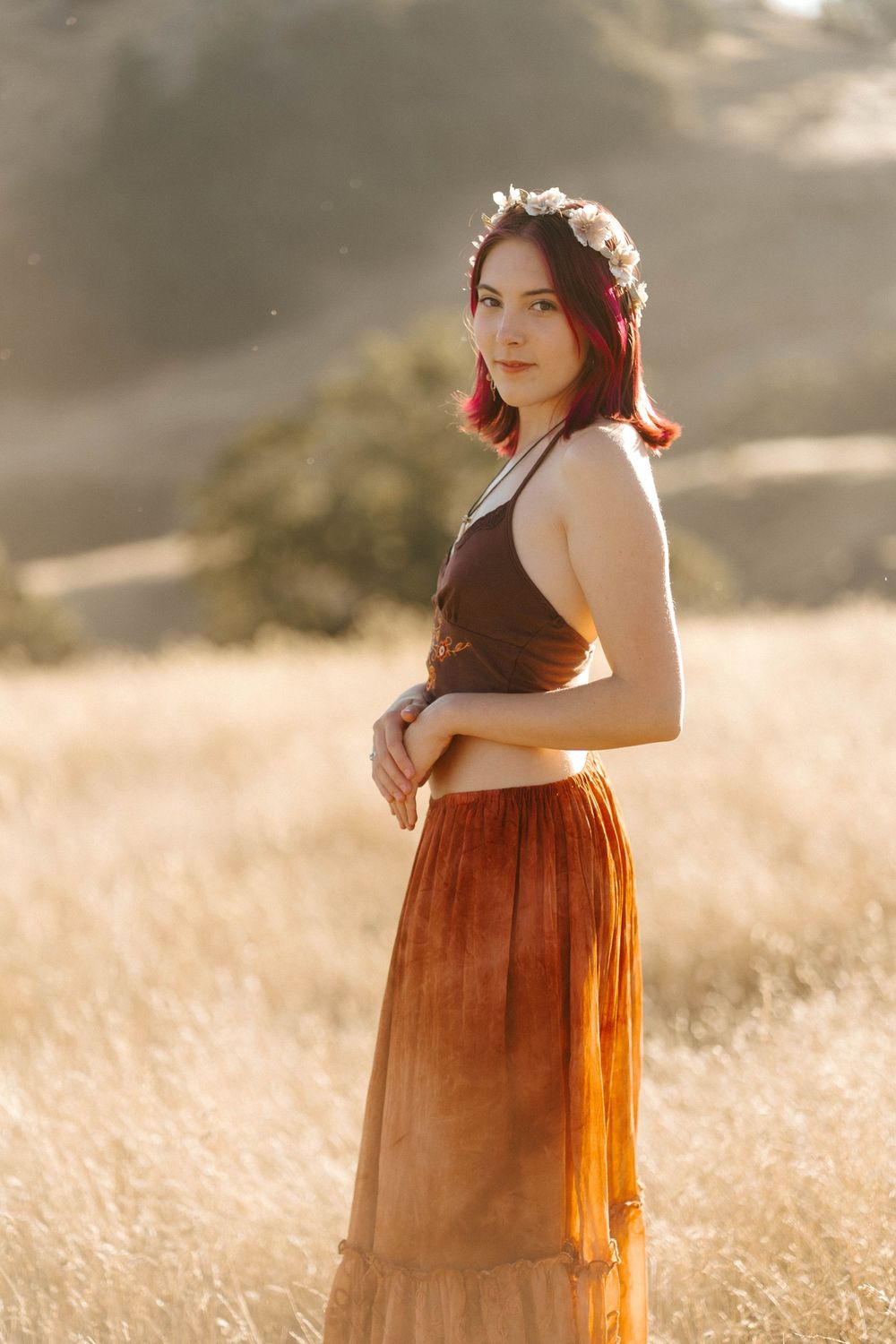 Young woman in brown crop top and rust maxi skirt with floral crown stands in golden field at sunset.