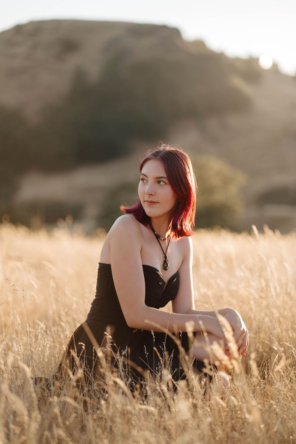 Young woman with red hair in black dress sitting in golden field at sunset with hills in background.