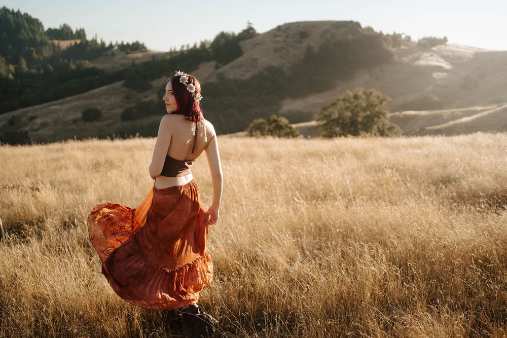 Woman in flowing rust-colored bohemian skirt standing in golden field with sunlit hills in background.