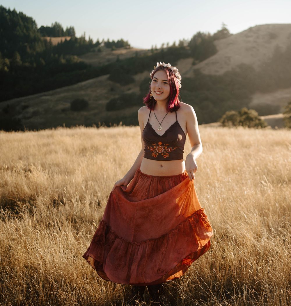 Young woman with pink hair wearing a floral crown, dark crop top, and rust tiered maxi skirt twirling in a golden meadow.