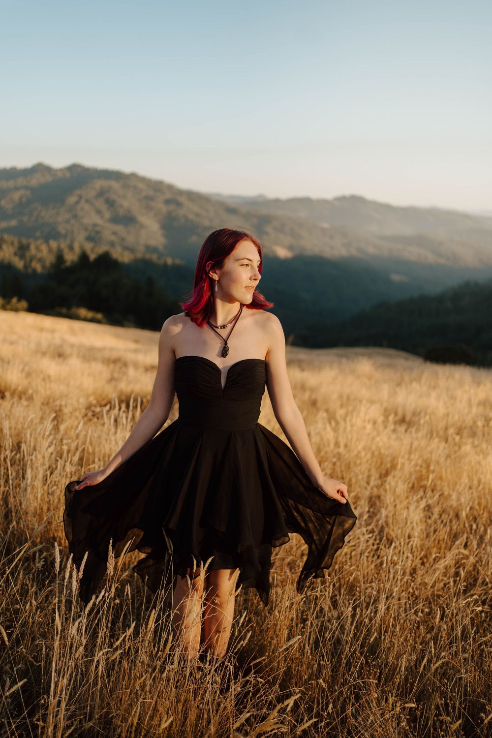 Young woman with red hair in black strapless dress and boots standing in golden field with mountain backdrop.