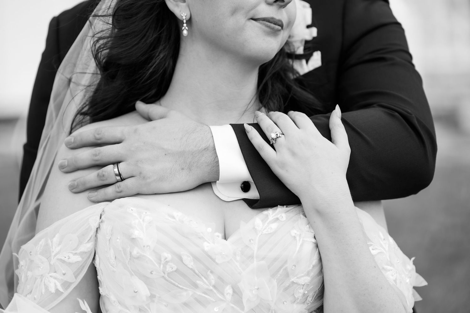 Black and white wedding photo of bride and groom embracing, showcasing wedding rings and elegant attire.