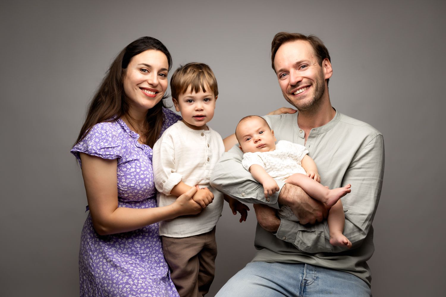 Happy family portrait with parents and two young children, including a baby, posing together against a gray background.