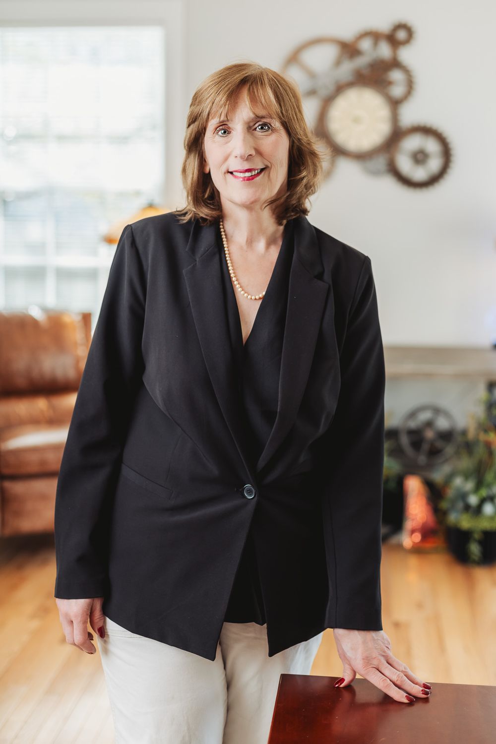 Professional woman in black blazer and pearl necklace standing confidently in a modern office setting.