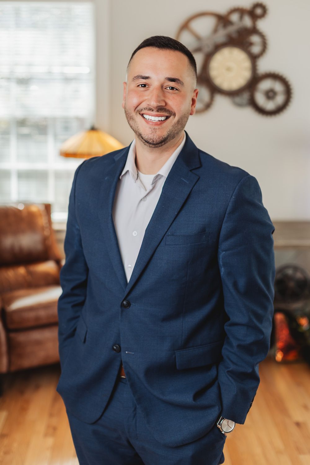 Smiling man in navy blue suit with hands in pockets standing in a modern living room with gear wall decor.