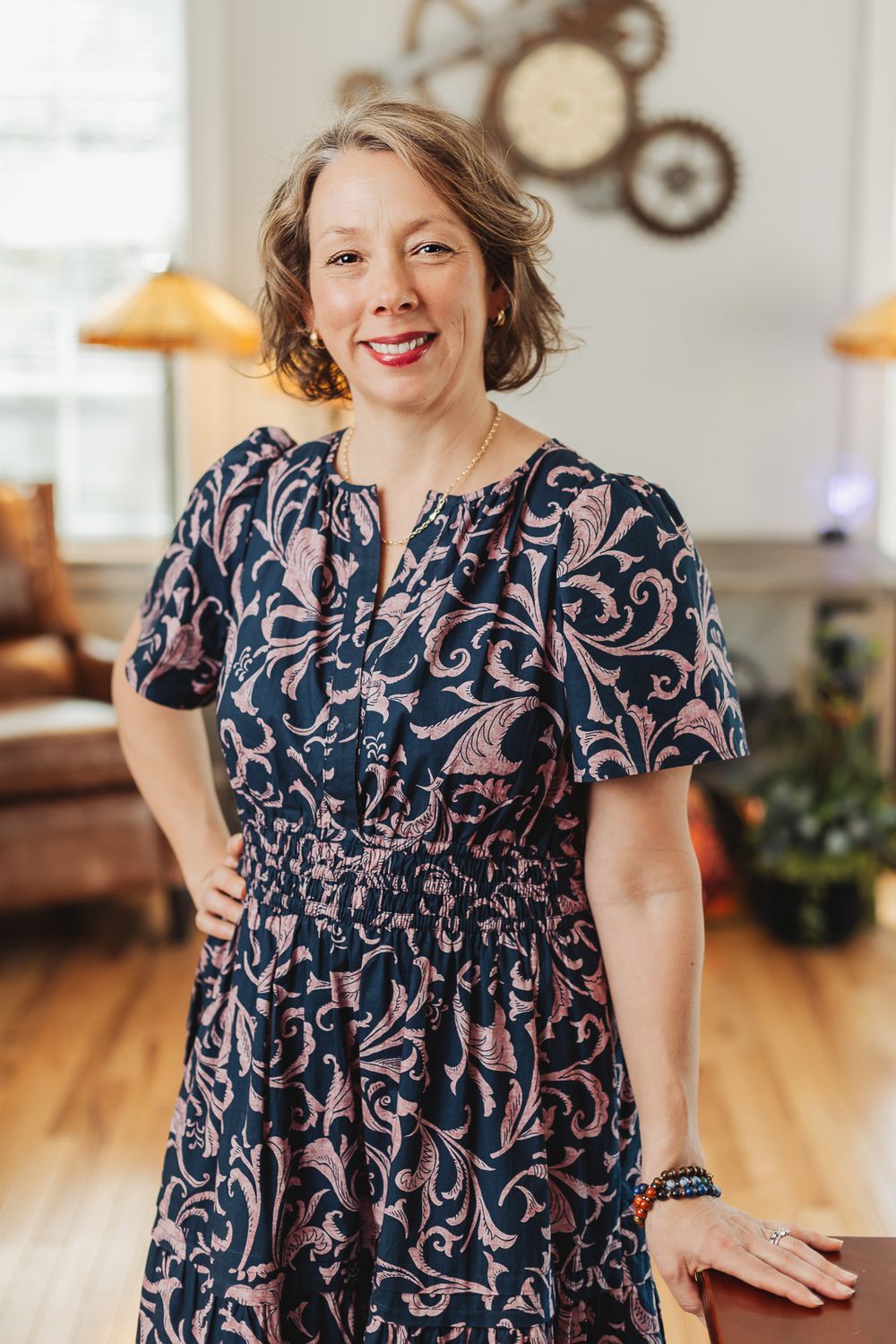 Woman in navy floral dress smiling confidently in a warmly decorated living room interior.