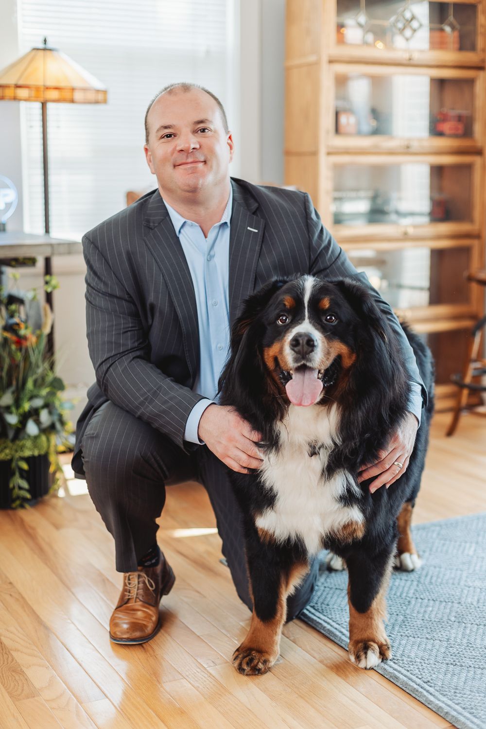 Man in suit crouching beside a Bernese Mountain Dog on hardwood floor in a warmly lit home interior.