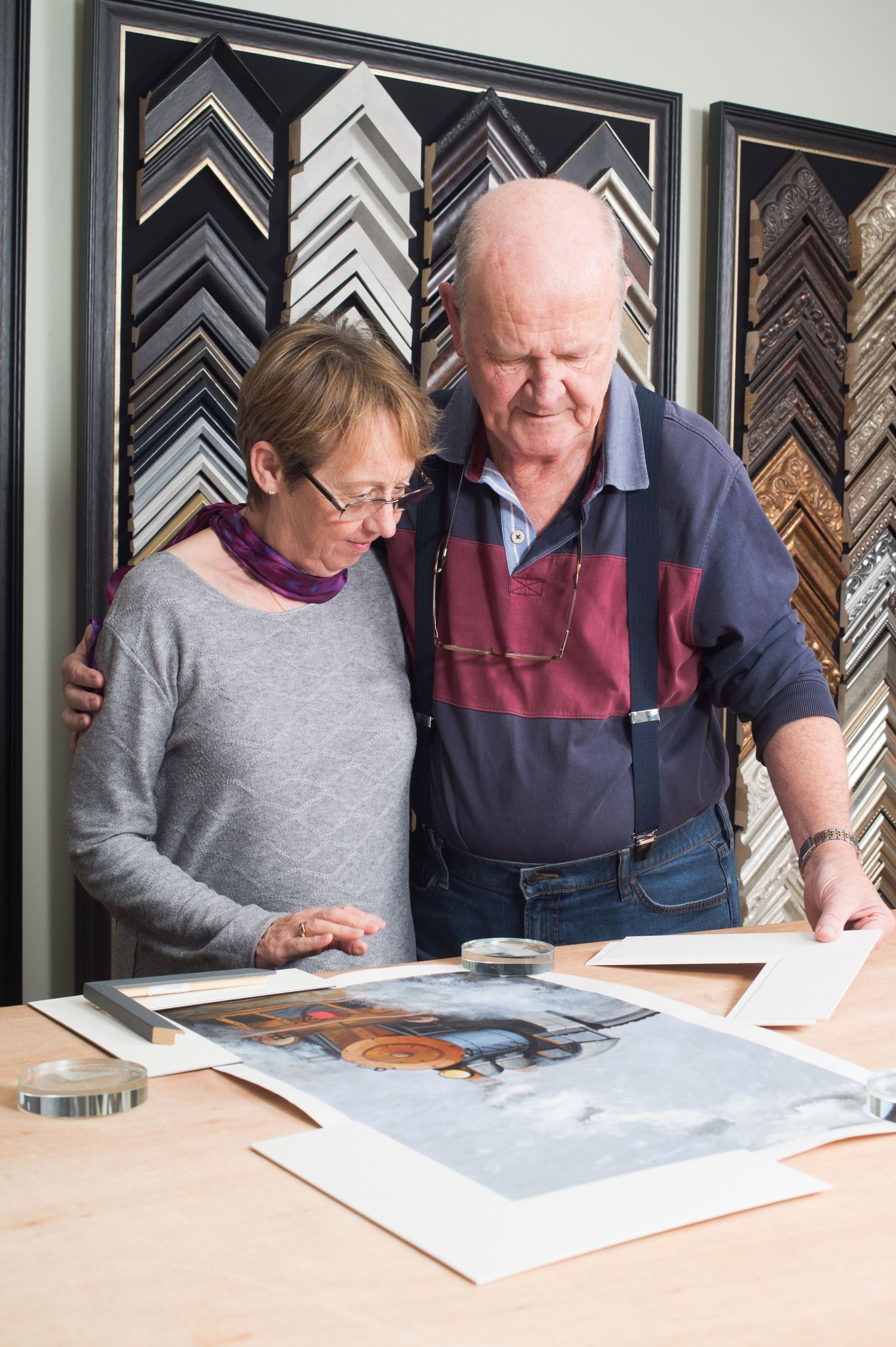 An elderly man and woman stand closely, selecting frames for an artwork in a frame shop. They appear thoughtful, surrounded by a display of