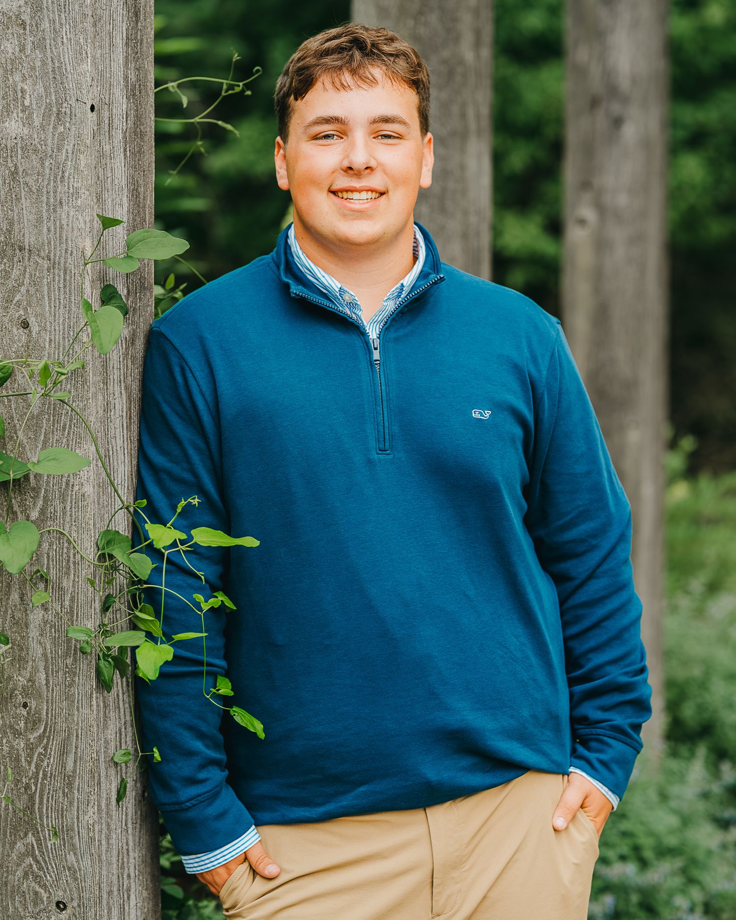 Young man in blue suit and red tie leaning against tree trunk outdoors, smiling for formal portrait photo.