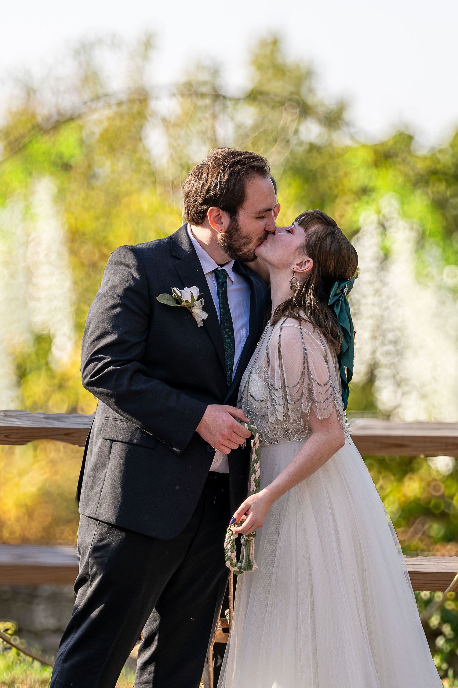 A bride and groom share their first kiss on a wooden bridge surrounded by lush greenery at an outdoor wedding ceremony.