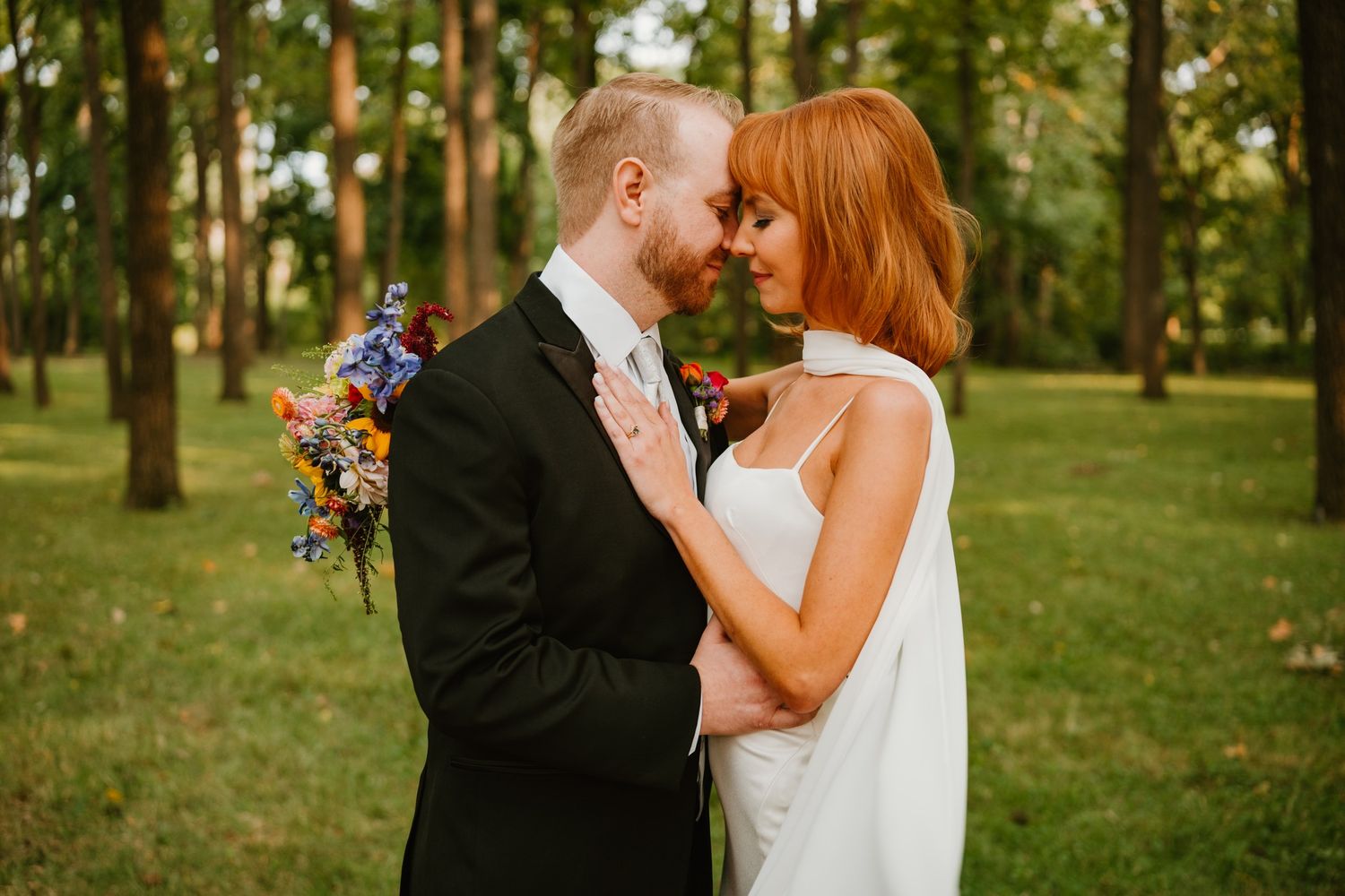 Bride and groom share a romantic forehead touch outdoors, surrounded by lush green trees, holding a colorful wildflower bouquet.