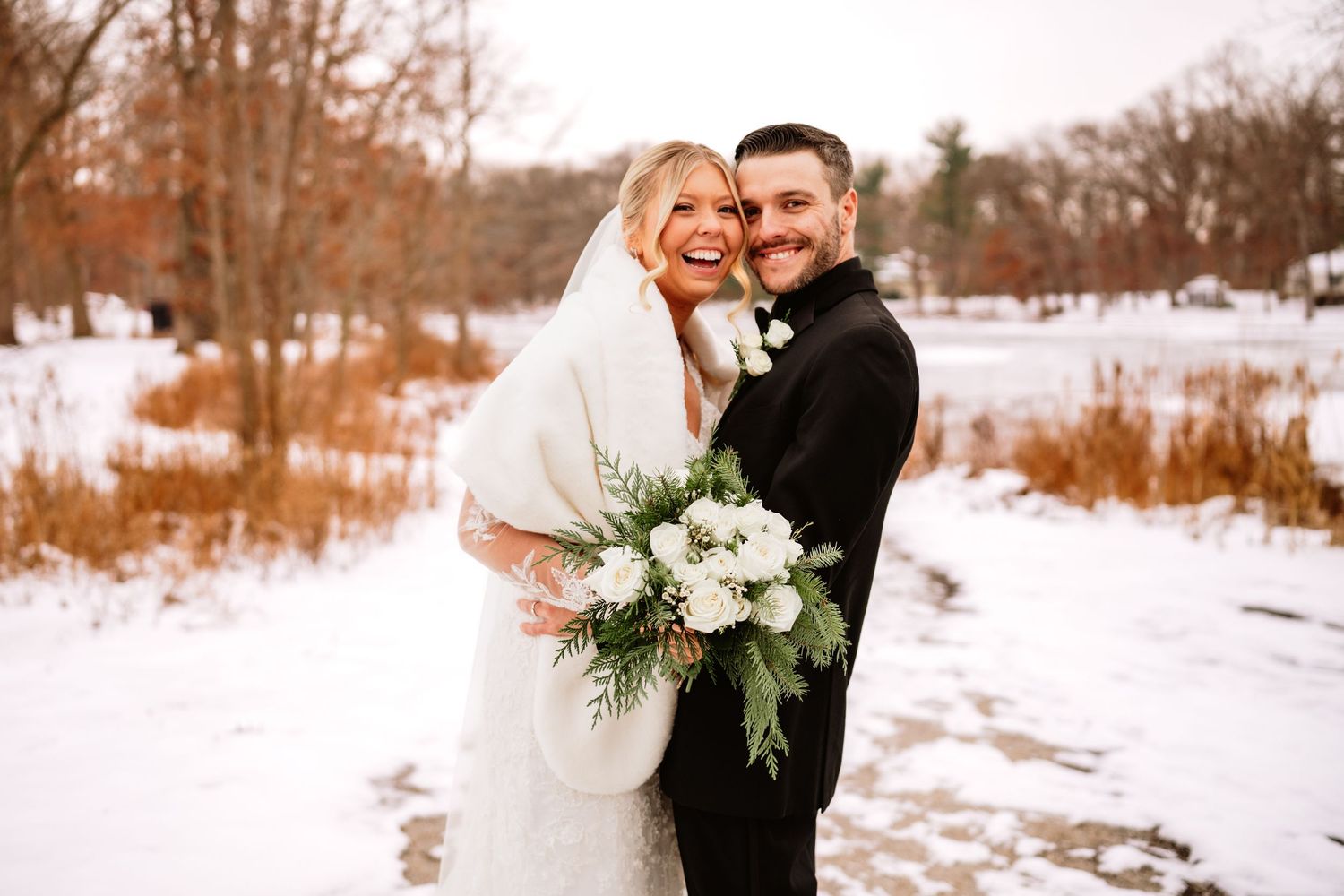 Bride and groom smiling together in snowy winter outdoor wedding portrait, bride holding white floral bouquet with greenery.