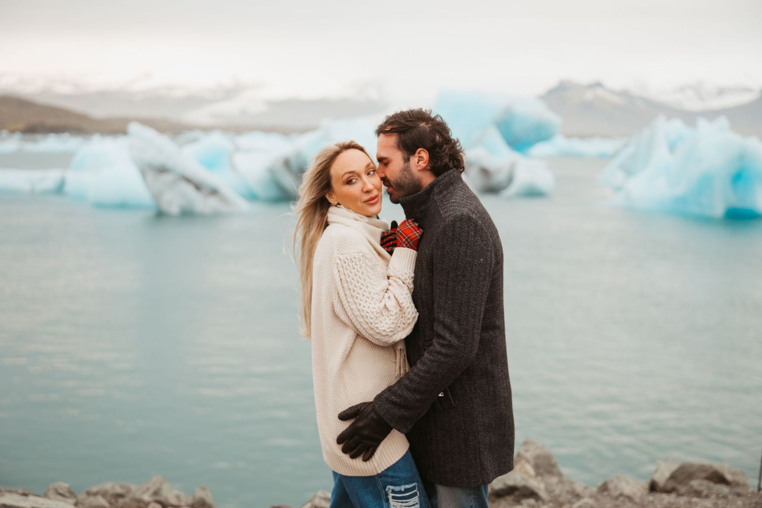 Couple embracing near glacial lagoon with blue icebergs in Iceland, woman in cream sweater and man in gray knit jacket.