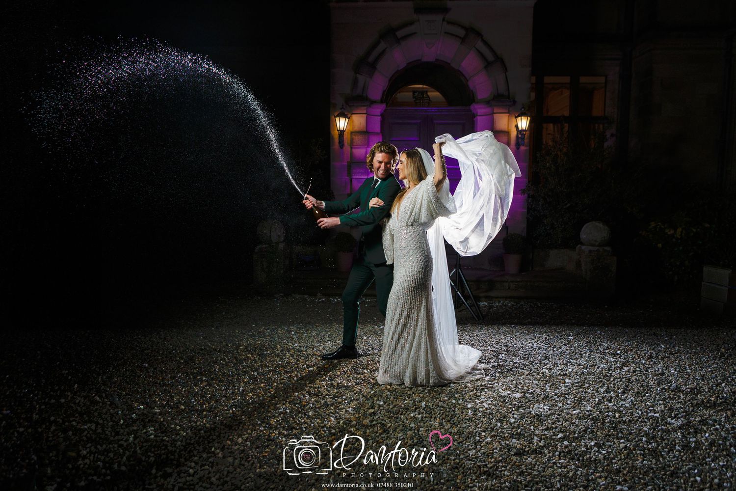 Bride and groom celebrating with champagne spray at night outside elegant venue with purple lighting.