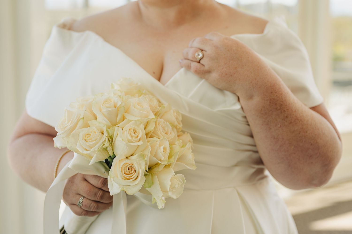 Bride in white off-shoulder gown holding bouquet of cream roses, wearing diamond ring.