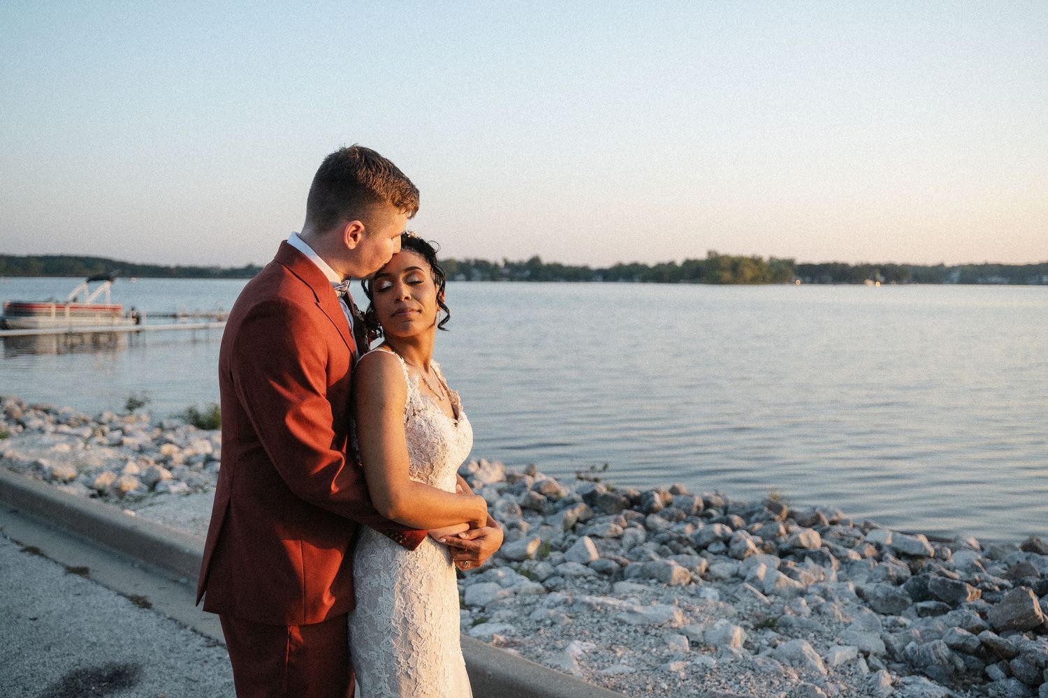 Black and white photo of a couple in wedding attire embracing by a calm lake at dusk.