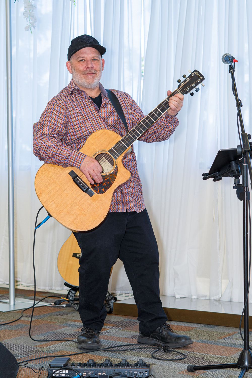 Middle-aged man playing acoustic guitar on stage, wearing black cap and plaid shirt, performing at an indoor event.