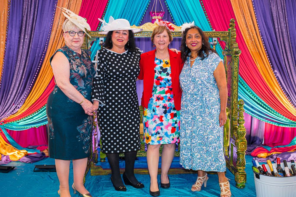 Four women in colorful dresses pose together in front of vibrant draped fabric backdrop at a festive event.