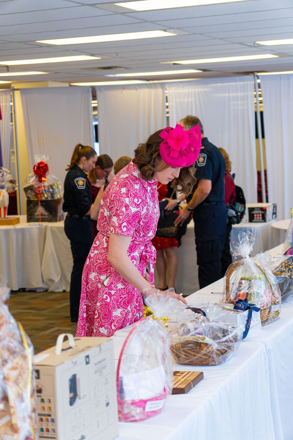 Woman in pink floral dress and hat browses gift baskets at a charity auction event with police officers in background.