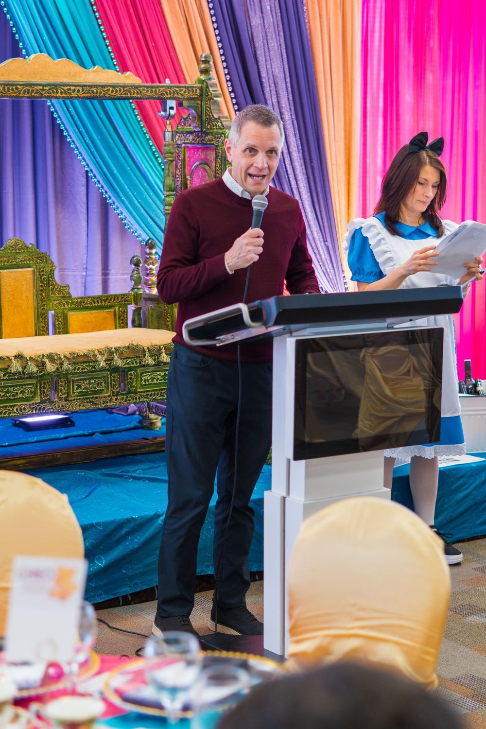 Man speaking at podium with microphone at colorful themed event with decorative drapes.