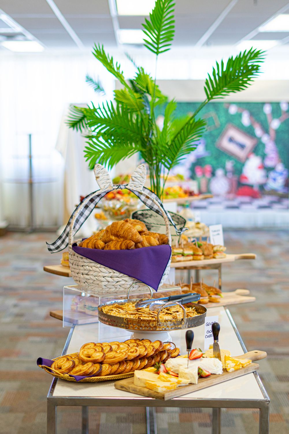 Elegant buffet table with croissants in a basket, crackers, cheese, and pastries decorated with tropical palm leaves.