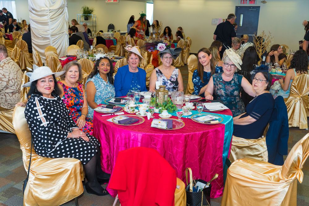 Women in decorative hats gather around a pink tablecloth table at an elegant banquet hall celebration.
