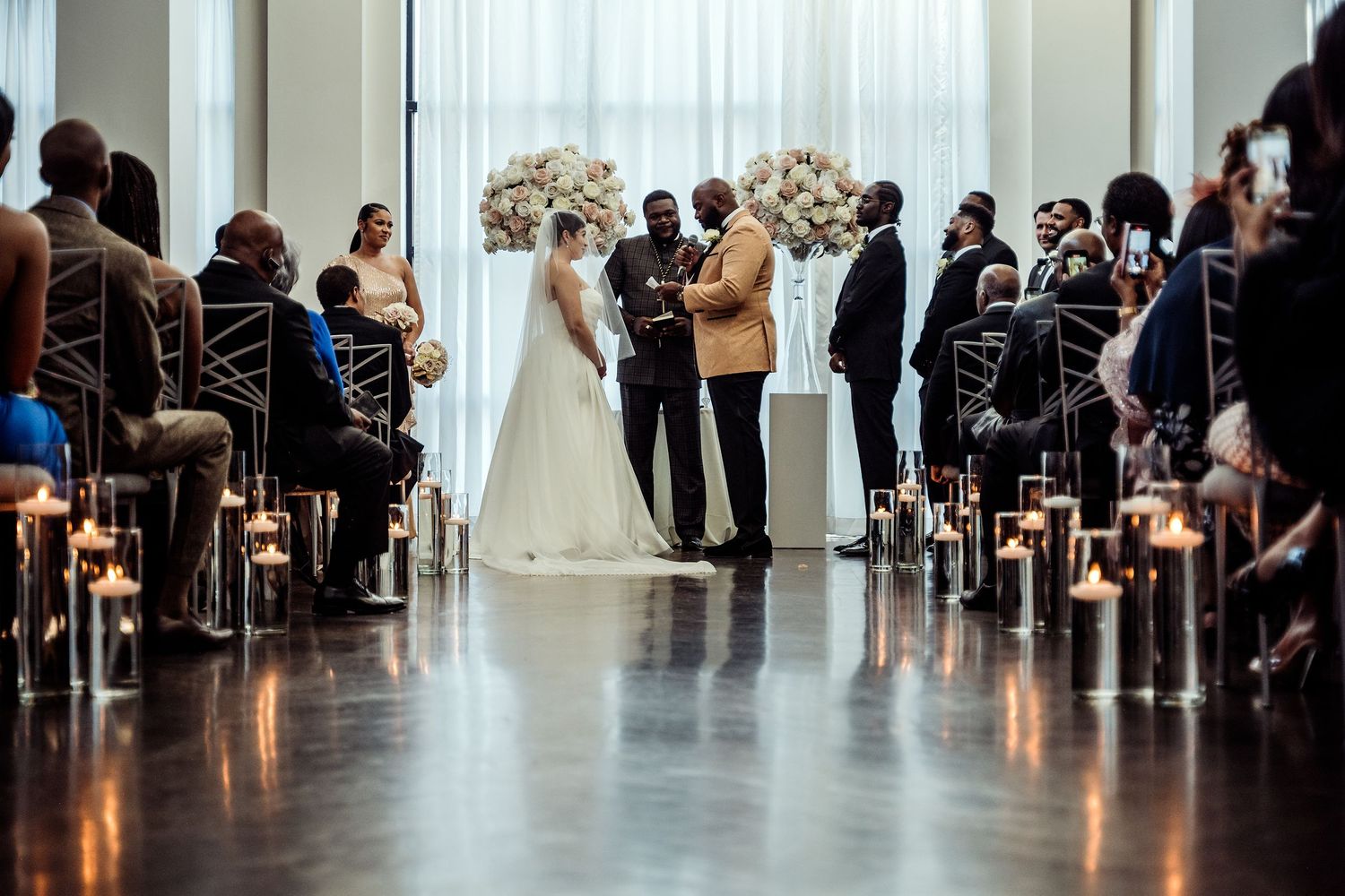 Elegant indoor wedding ceremony with candle-lined aisle, floral arrangements, and guests seated in a modern venue.