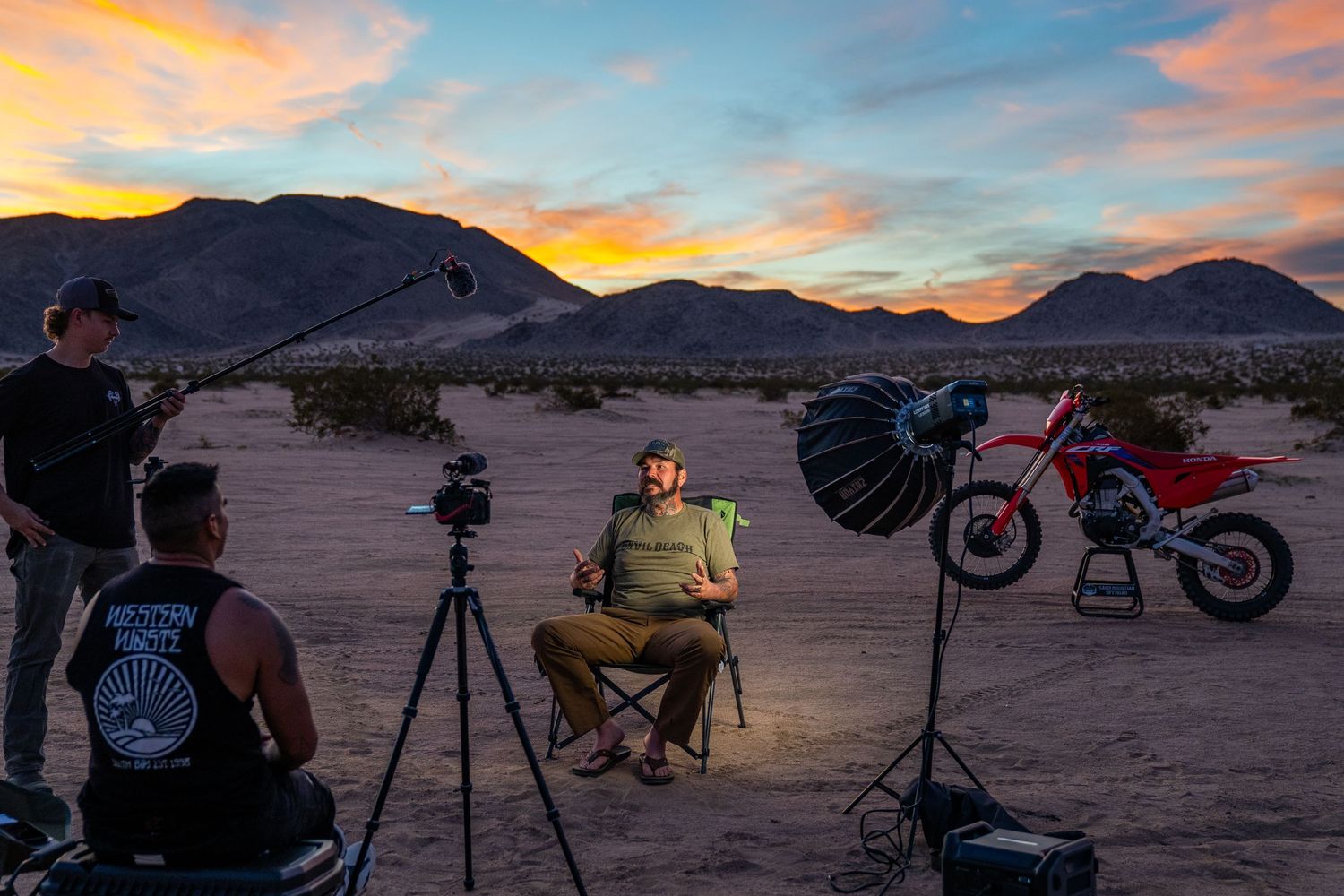 Man seated in desert with camera tripod and lighting equipment next to a red dirt bike, mountains in background.