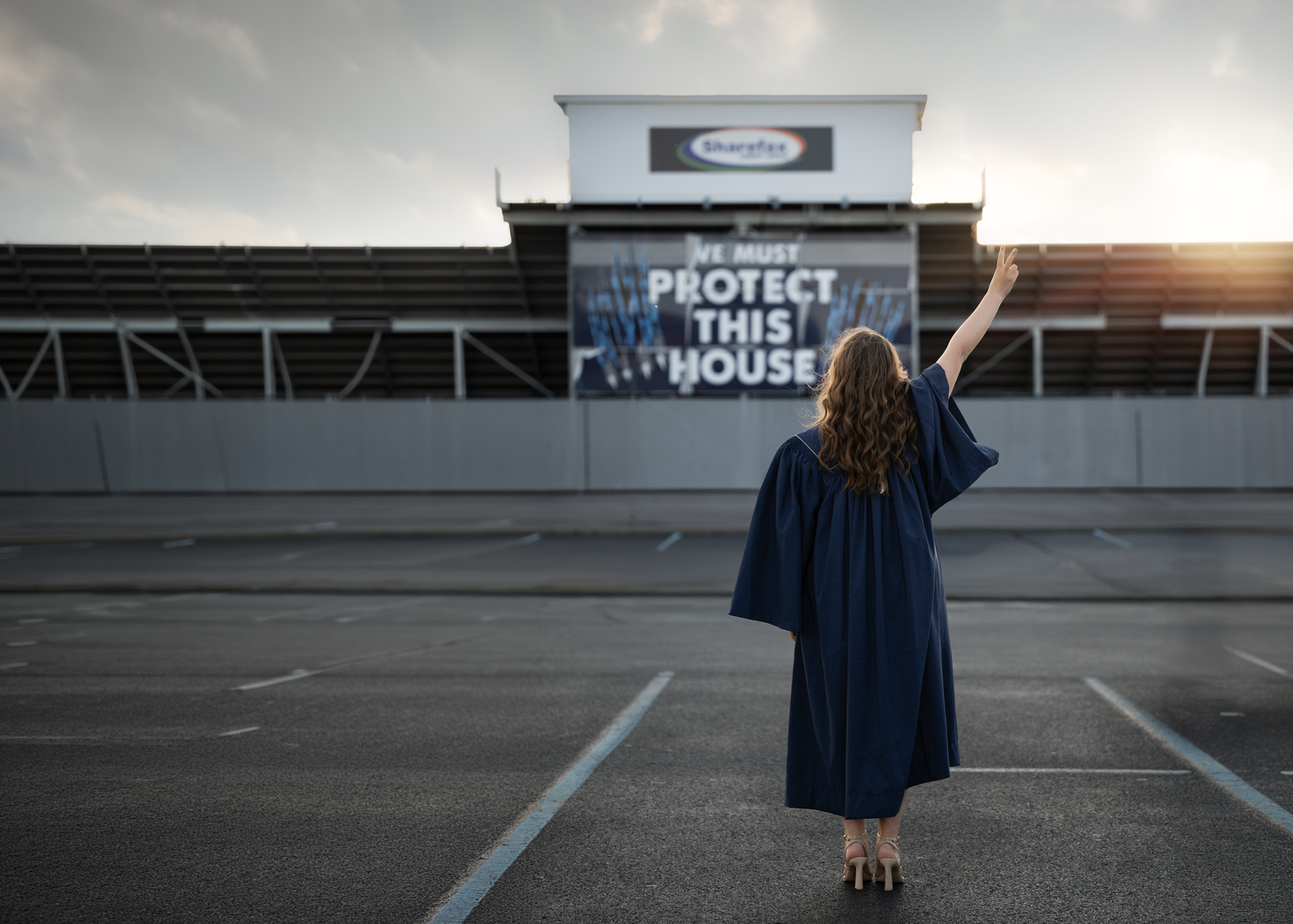 Graduate in cap and gown raises arms before a stadium sign reading Protect This House at sunset.