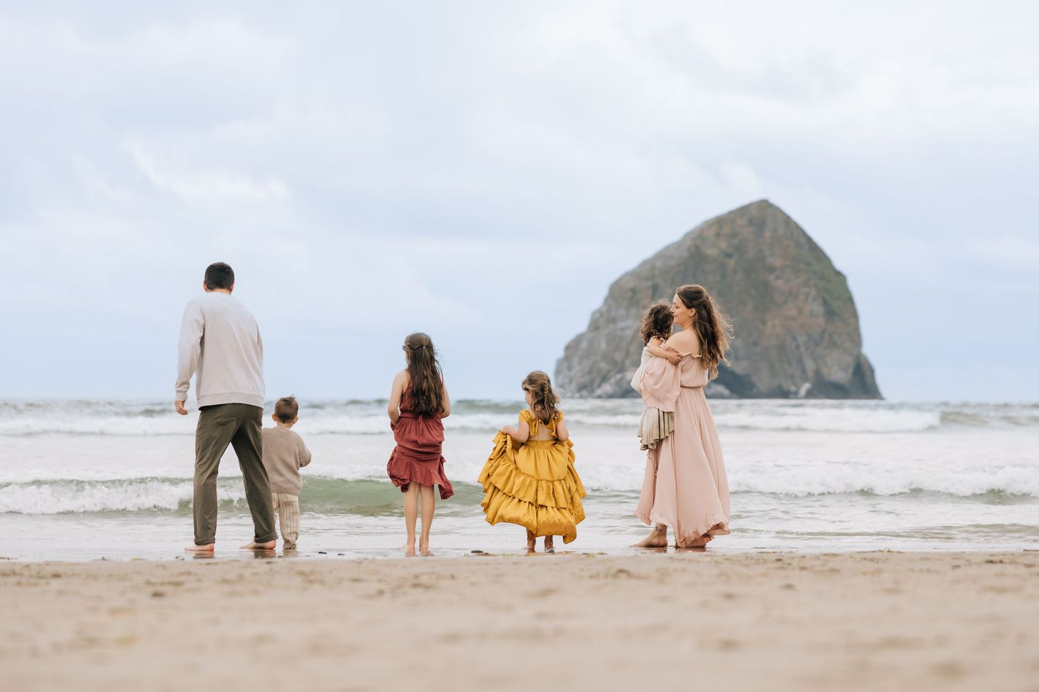 Family of five standing on a sandy beach gazing at Haystack Rock at Cannon Beach, Oregon on a cloudy day.