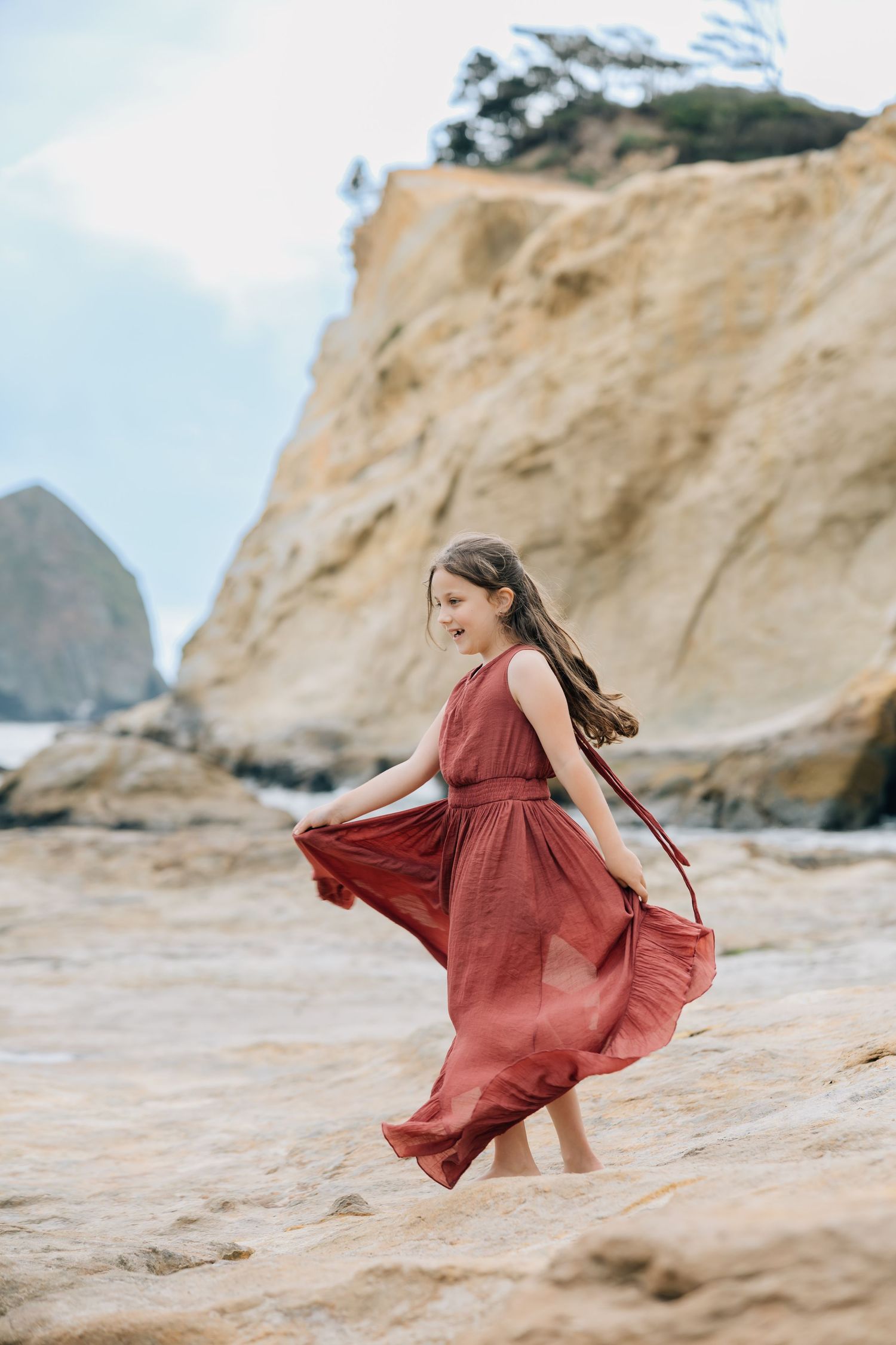 A young girl in a flowing red dress twirls on rocky beach with dramatic coastal cliffs in the background.