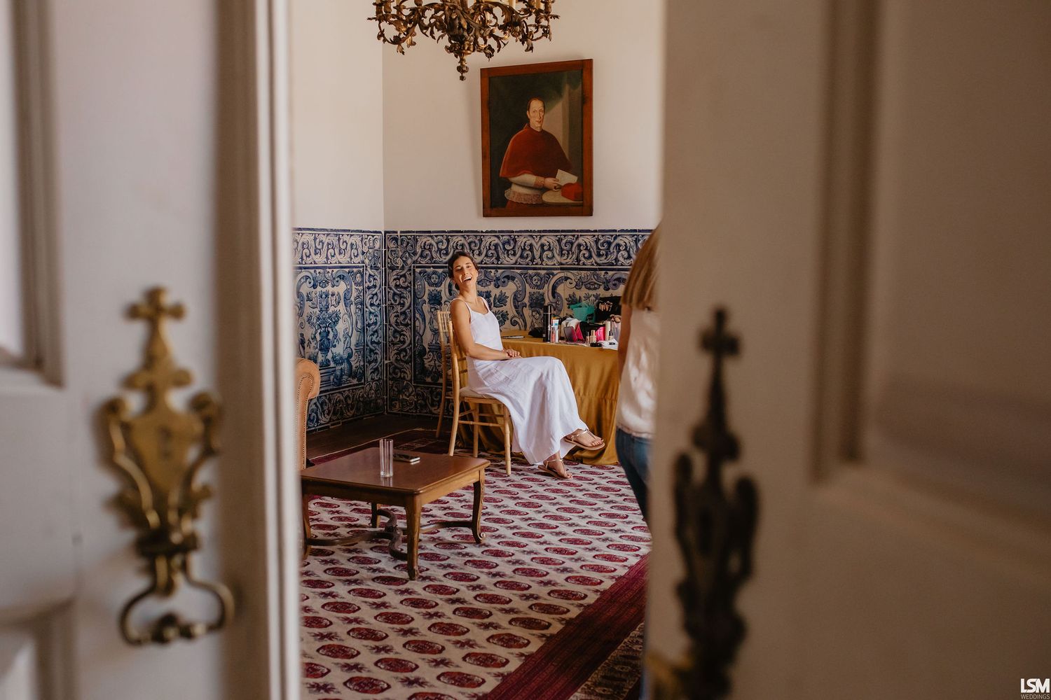 Bride in white dress sitting in ornate room with blue azulejo tiles and portrait painting, elegant wedding preparation.