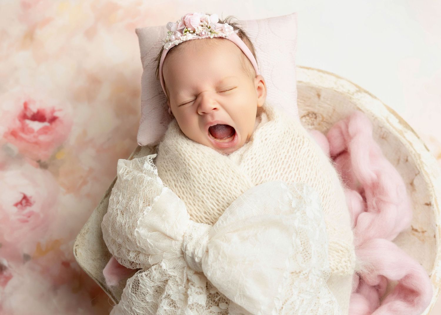 Adorable newborn baby girl yawning, wrapped in white knit blanket with floral headband, resting on pink background.