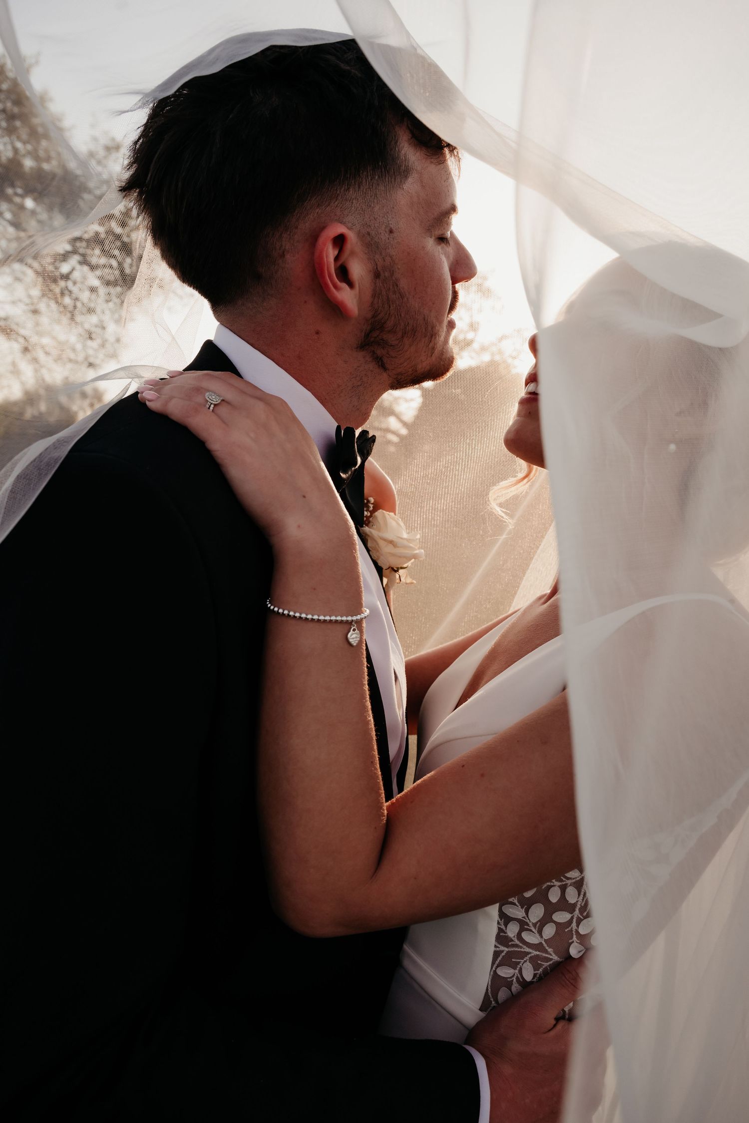A groom in a black suit embraces his bride in a white dress and veil during an outdoor wedding ceremony.