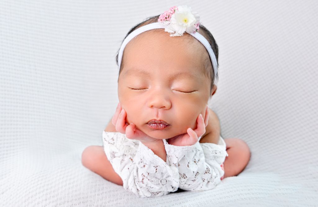 Sleeping newborn baby girl wearing white lace outfit and floral headband, posed with hands under chin. Newborn Photographer in Cypress TX