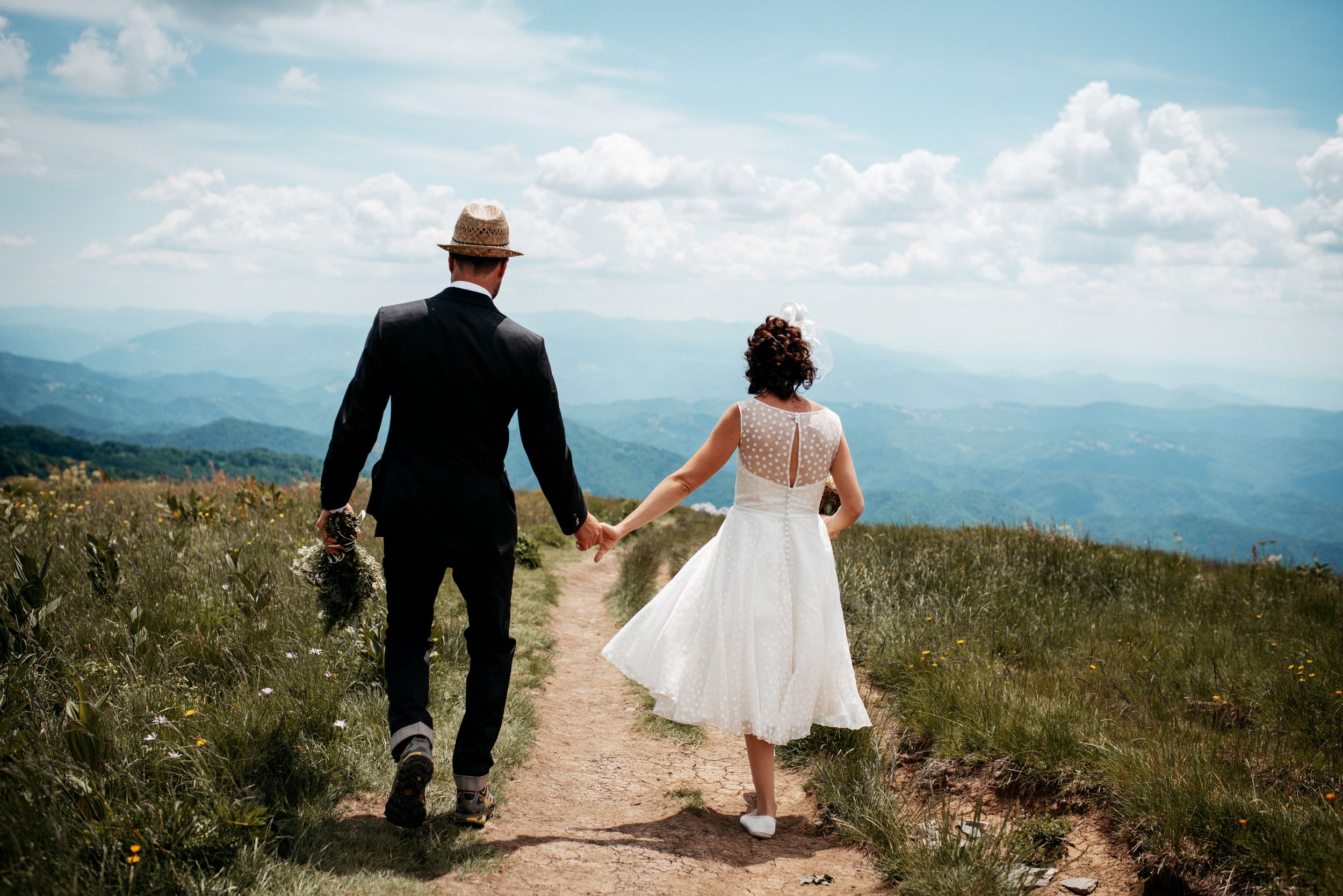 Couple walking hand in hand on mountain trail with scenic mountain vista in background.