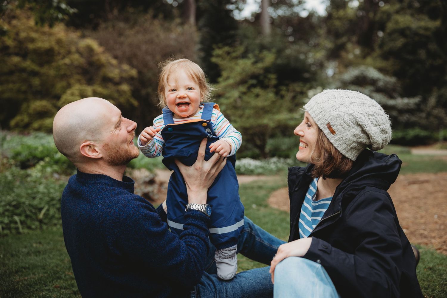 Happy family with bald father and mother in knit hat lifting laughing toddler outdoors in lush green park.