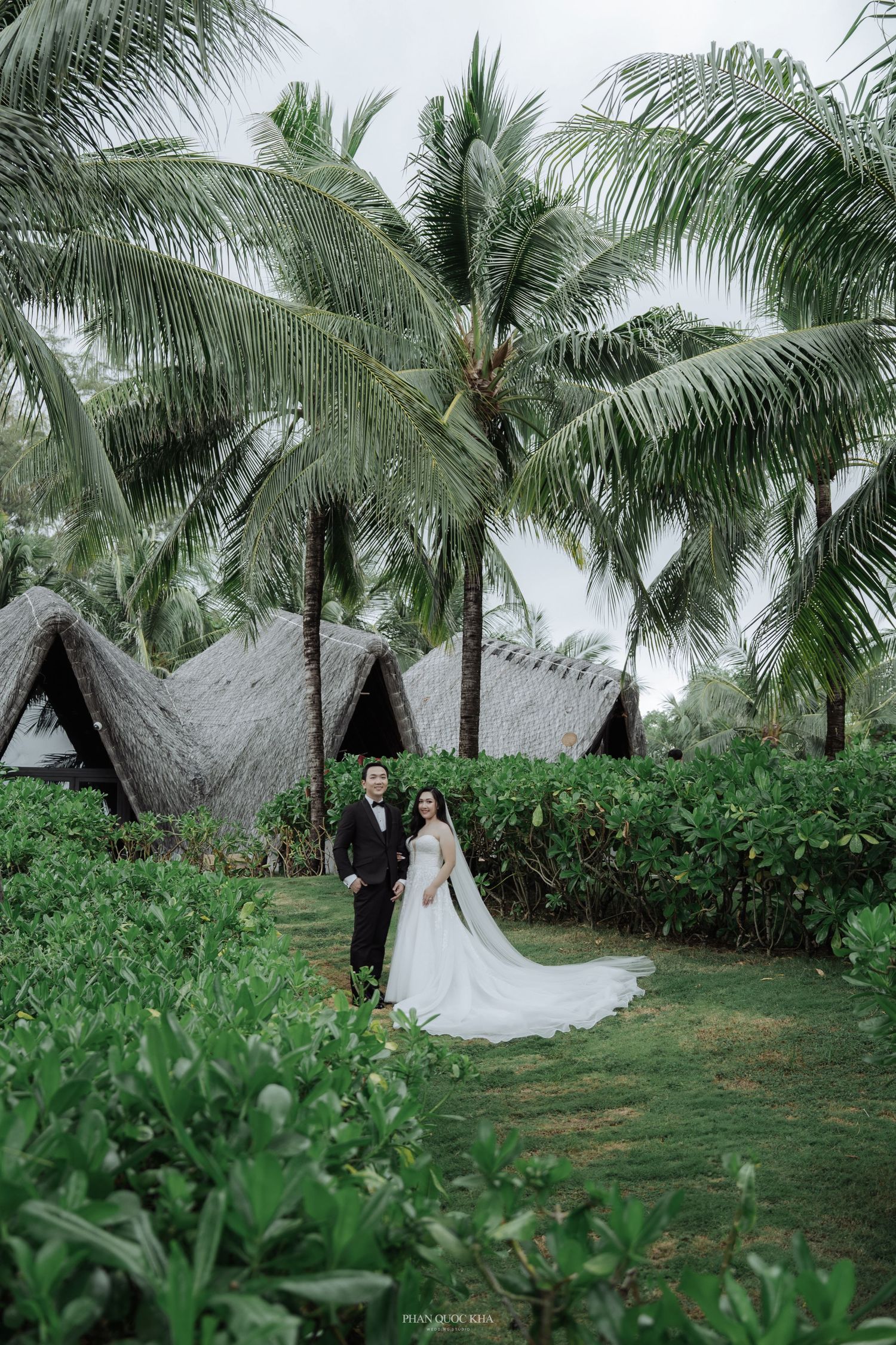Elegant bride and groom posing among tropical palm trees and thatched roof bungalows.