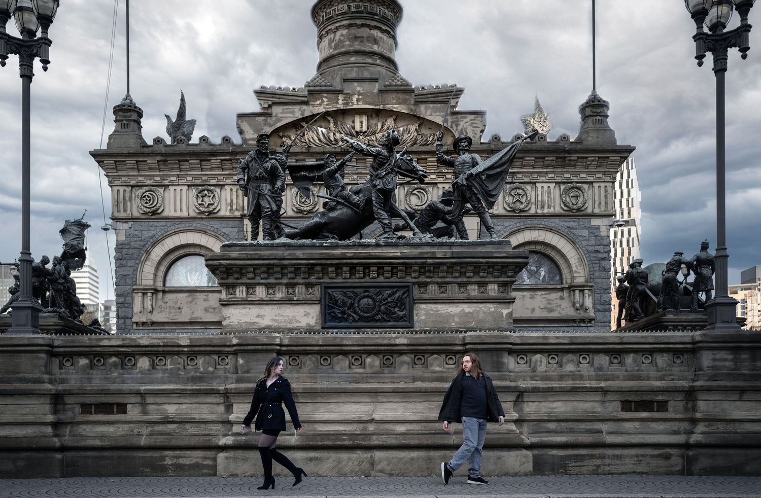 Two people walking in front of a grand ornate monument with sculptures under a cloudy sky.
