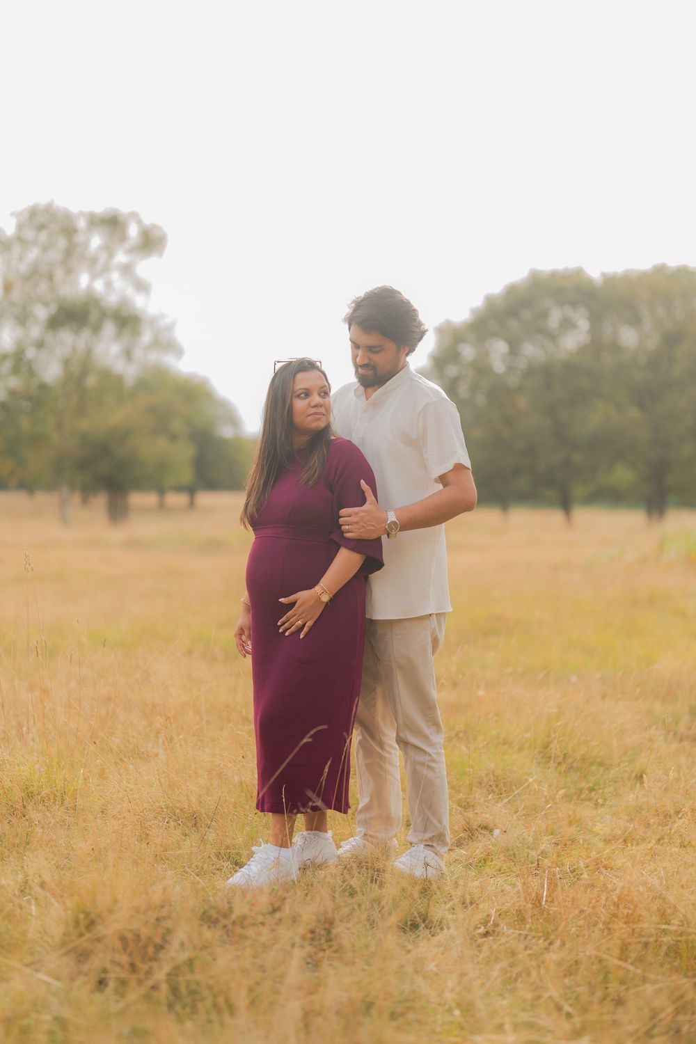 Couple in a golden field, pregnant woman in burgundy dress embraced by partner in neutral tones.