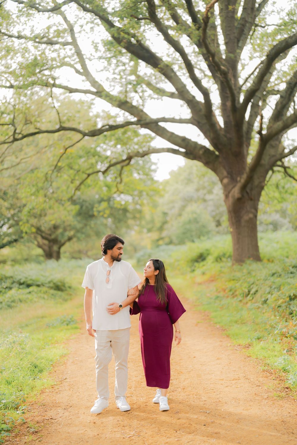 Couple walking on a dirt path surrounded by large trees, woman in burgundy dress and man in white outfit.