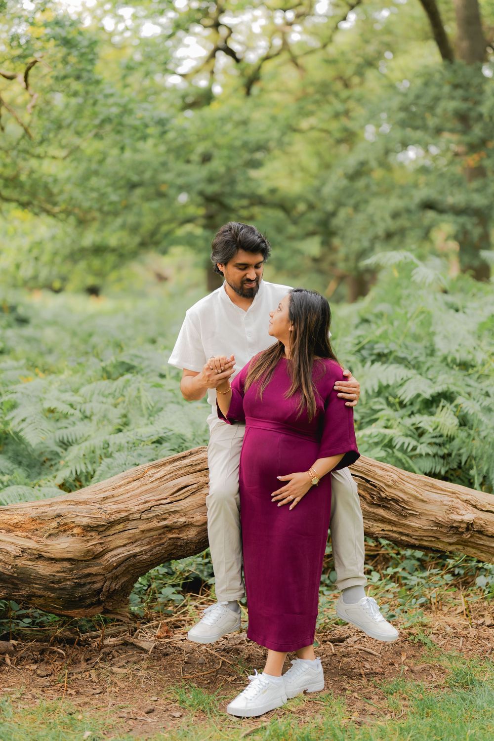 Pregnant woman in magenta dress with partner on log in lush green forest setting.