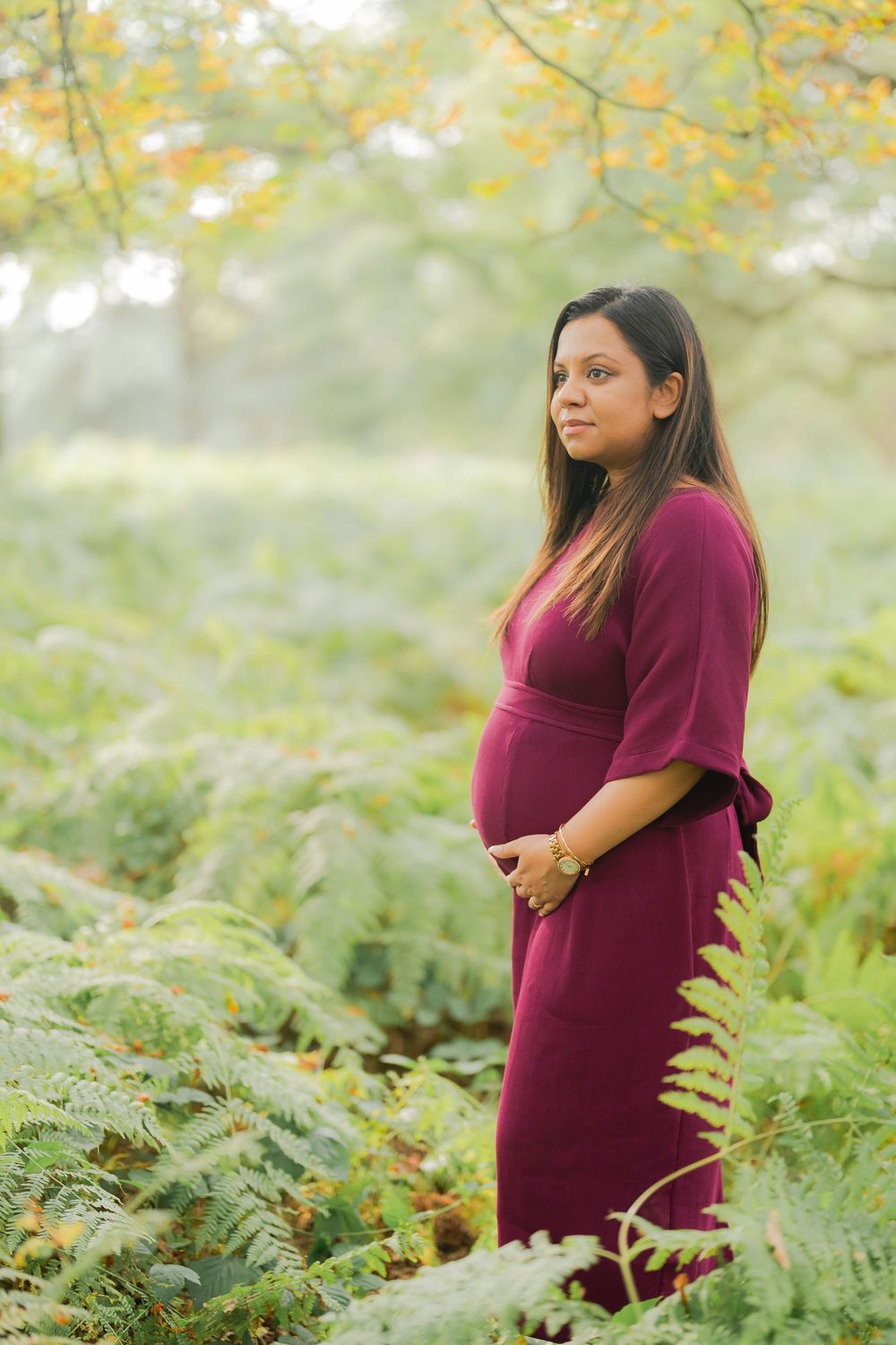 Pregnant woman in burgundy dress standing among green ferns in a misty autumn forest, cradling her baby bump.