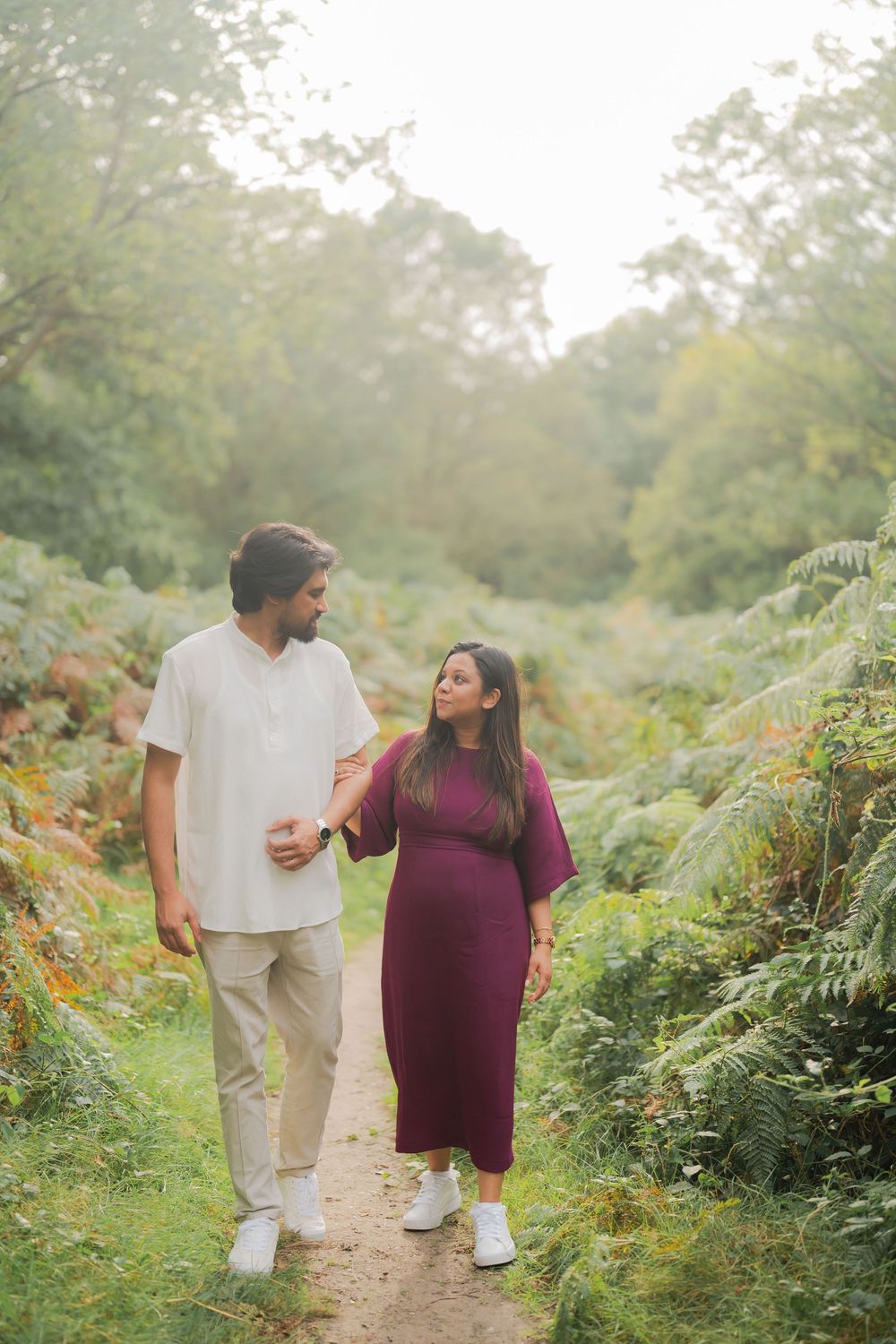 Pregnant couple walking on a garden path surrounded by lush greenery, woman in burgundy dress, man in white shirt.