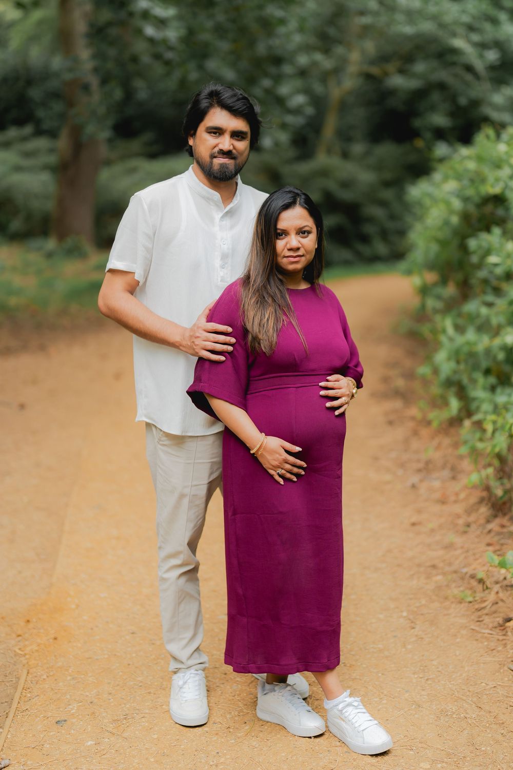 A pregnant woman in a purple dress and her partner pose together on a garden path surrounded by lush greenery.