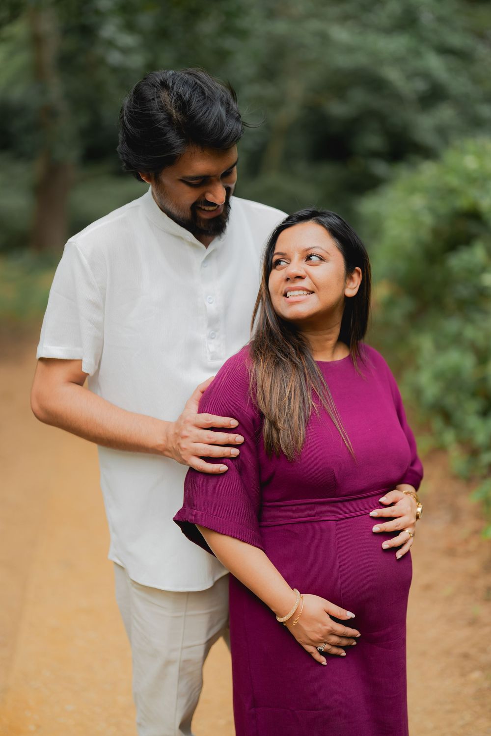 Expecting couple poses outdoors on dirt path, man in white shirt embraces pregnant woman in magenta dress, lush green trees behind.
