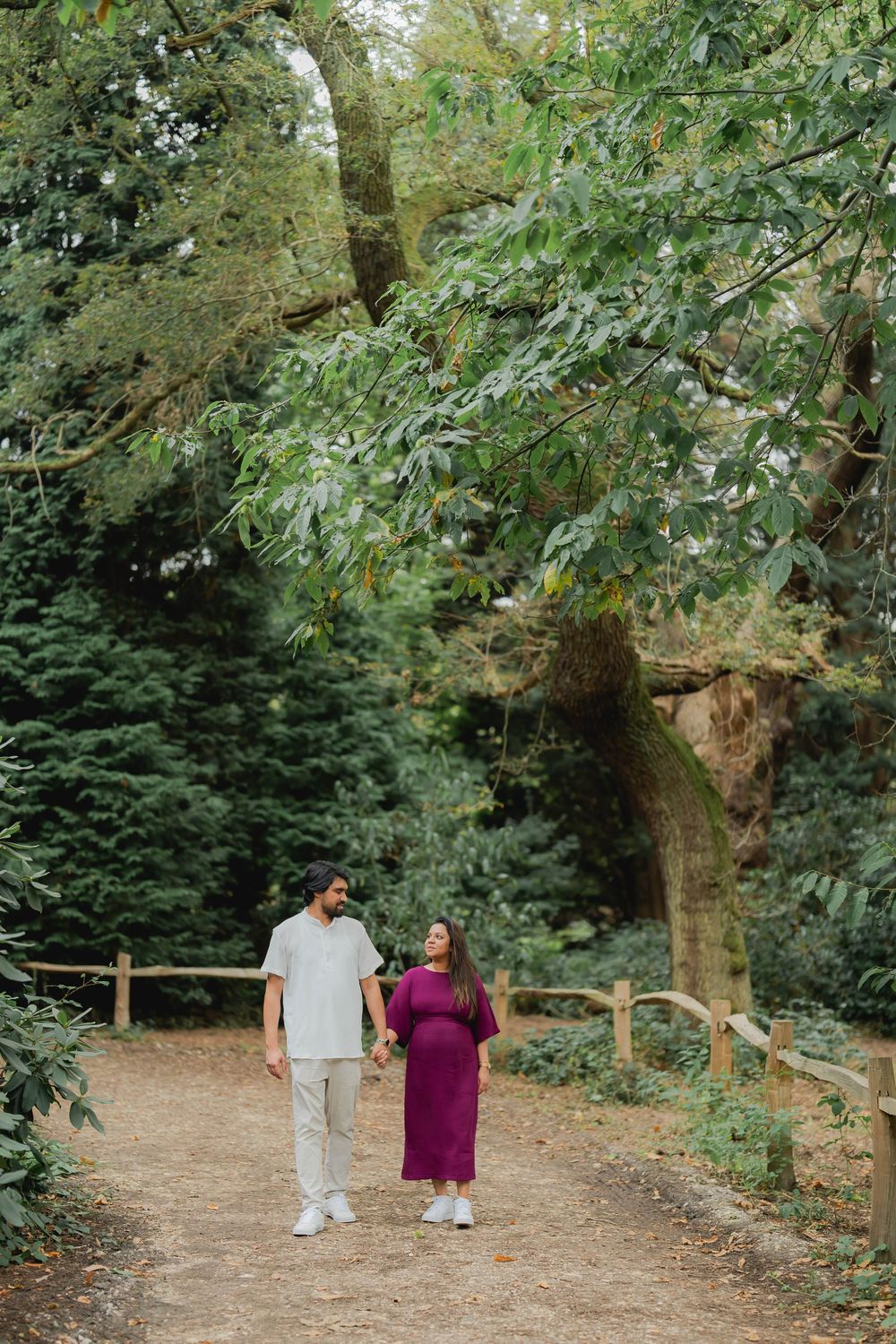 Pregnant couple walking hand in hand on a woodland path surrounded by lush green trees.