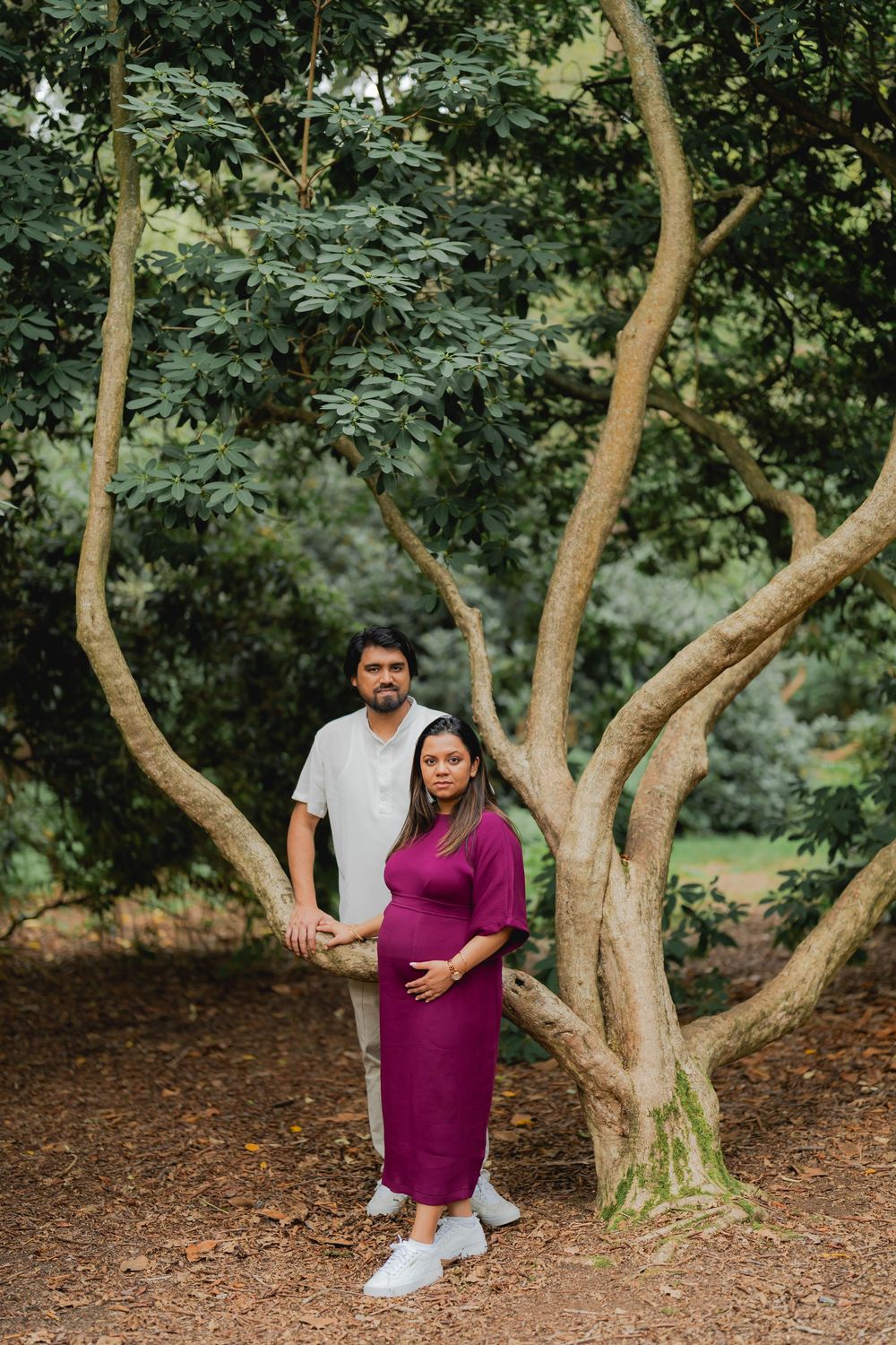 Pregnant couple posing under a large tree in a lush green park, woman in magenta dress cradling her baby bump.