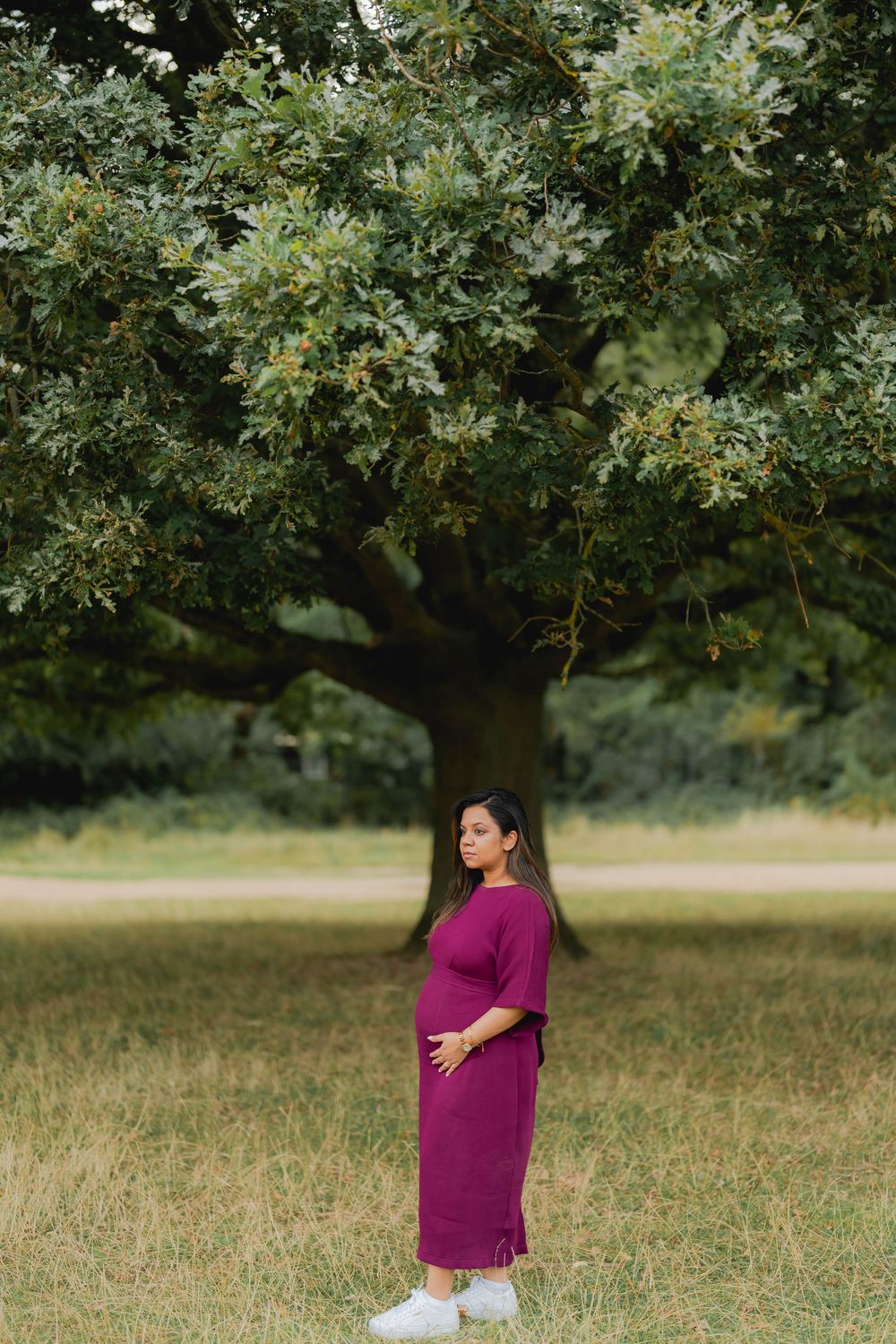 Pregnant woman in purple dress standing under large oak tree in park, cradling baby bump outdoors.